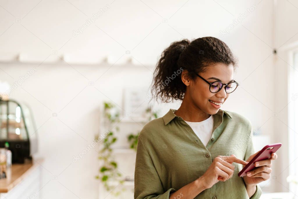 Young black woman in eyeglasses smiling while using cellphone at cafe indoors