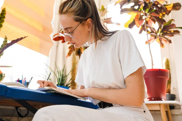 眼鏡をかけながらノートを書きながら屋内で働く若い白人女性 — ストック写真