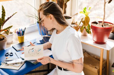 Genç beyaz kadın evde çalışırken çizimler yapıyor.