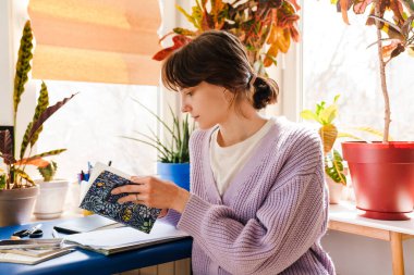 Genç beyaz kadın evde otururken eskiz defterine bakıyor.