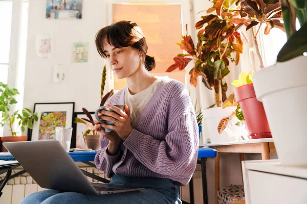 屋内でノートパソコンを操作しながらコーヒーを飲む若い白人女性 — ストック写真