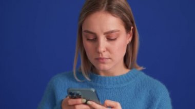 Mavi kazak giyen neşeli kadın mavi stüdyoda telefonla mesajlaşıyor.