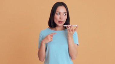 Mavi tişörtlü konsantre bir kadın bej renkli stüdyoda telefonla bir mesaj kaydediyor.