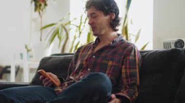 Ekose gömlekli gülümseyen yetişkin bir adam telefonla mesaj atıyor ve evde konuşuyor.