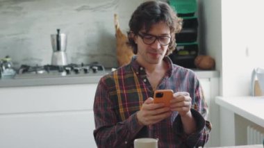 Ekose gömlekli yakışıklı yetişkin adam mutfakta telefonla mesaj atıyor.