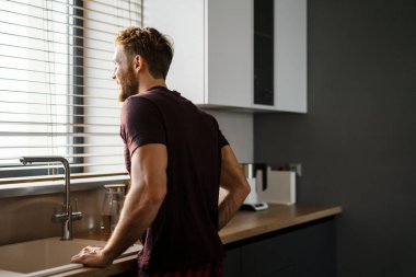 Mutfak lavabosunun önünde dikilirken pencereden bakan çekici, mutlu genç adam.