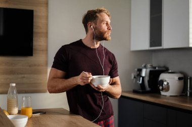 Çekici, sağlıklı bir genç adam mutfakta elinde kâseyle durup kulaklıkla müzik dinlerken lezzetli bir kahvaltı yapıyor.