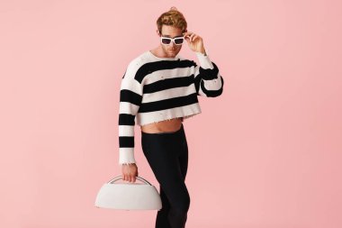 Kızıl saçlı, güneş gözlüklü genç adam pembe duvarda izole edilmiş halterli poz veriyor.