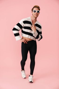 Güneş gözlüklü genç adam gülümsüyor ve pembe duvarın üzerinden izole edilmiş kameraya bakıyor.