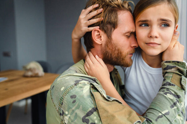 Мужской грустный военный обнимает свою расстроенную дочь в помещении