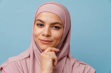 Tesettürlü genç Müslüman kadın gülümsüyor ve mavi arka planda izole edilmiş kameraya bakıyor.