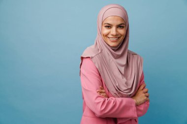 Tesettürlü genç Müslüman kadın gülümsüyor ve mavi arka planda izole edilmiş kameraya bakıyor.