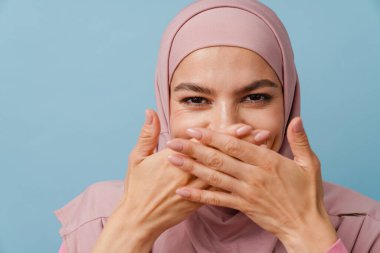 Genç Müslüman kadın, mavi arka planda ağzını kapatırken kameraya bakıyor.