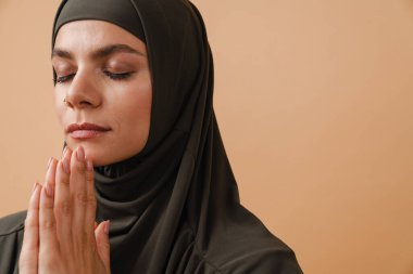 Tesettürlü genç Müslüman kadın bej arka planda izole edilmiş dua hareketi yapıyor ve poz veriyor.