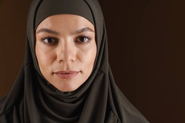 Tesettürlü genç Müslüman kadın kahverengi arka planda izole edilmiş kameraya bakıyor ve poz veriyor.