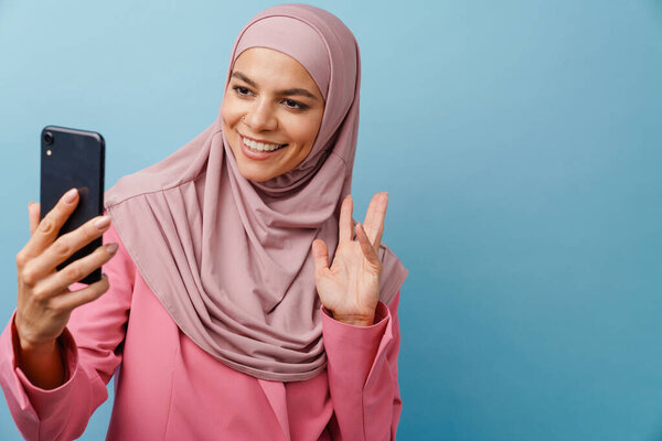 Young muslim woman in hijab gesturing while using mobile phone isolated over blue background