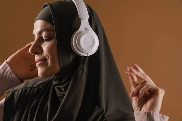 Nuori Musliminainen Hijabissa Kuuntelemassa Musiikkia Kuulokkeilla Eristetty Ruskealla Taustalla — kuvapankkivalokuva