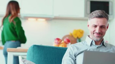 Dizüstü bilgisayarda çalışan ve karısıyla mutfakta konuşan olgun bir adam.