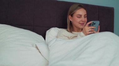 Gülümseyen sarışın kadın evde yatakta telefonla mesaj atıyor.