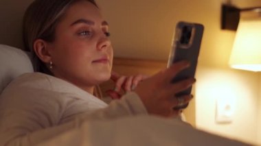 Düşünceli sarışın kadın evdeki yatakta telefonla mesaj atıyor.
