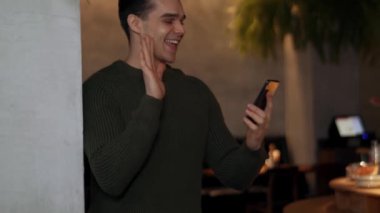 Süveter giyen neşeli bir adam kafede telefonla konuşuyor.