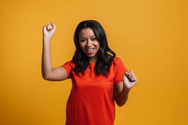 Mutlu orta yaşlı Afrikalı sıradan bir kadın sarı duvar arkasında duruyor, kutluyor, dans ediyor.