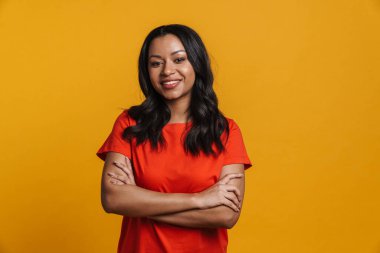 Orta yaşlı mutlu Afrikalı sıradan bir kadın sarı duvarın arkasında duruyor, kolları bağlı.