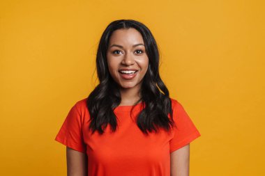 Genç siyah mutlu kadın gülümsüyor ve sarı arka planda izole edilmiş kameraya bakıyor.