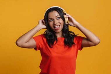 Sarı arka planda izole edilmiş kulaklıklarla müzik dinleyen siyahi genç bir kadın.