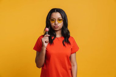 Güneş gözlüklü genç siyah kadın, sarı arka planda lolipop tutarken dudaklarını öpüyor.