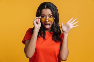 Güneş gözlüklü genç siyahi kadın sarı arka planda izole edilmiş bir şekilde el kol hareketi ve sürpriz yapıyor.