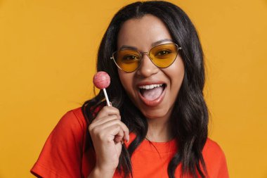 Sarı arka planda izole edilmiş lolipopla poz verirken gülümseyen güneş gözlüklü genç siyah kadın.