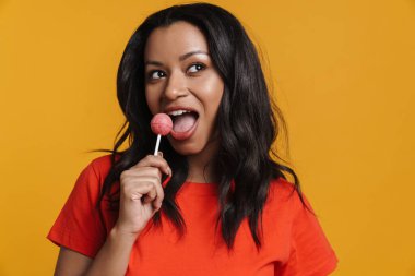 Sarı arka planda izole edilmiş lolipopu yalarken gülümseyen genç zenci kadın.