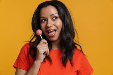Sarı arka planda izole edilmiş lolipopu yalarken gülümseyen genç zenci kadın.