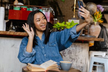 Gülümseyen Afrikalı genç bir kadın kafede otururken cep telefonuyla selfie çekiyor.