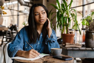 Kafe masasında otururken not defterine yazan düşünceli Afrikalı genç bir kadın.