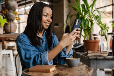 Kafede otururken cep telefonu kullanan gülümseyen Afrikalı bir kadın.