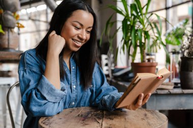 Kafe masasında otururken kitap okuyan gülümseyen Afrikalı genç bir kadın.