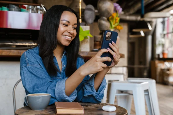 Black smiling woman using cellphone while sitting in cafe