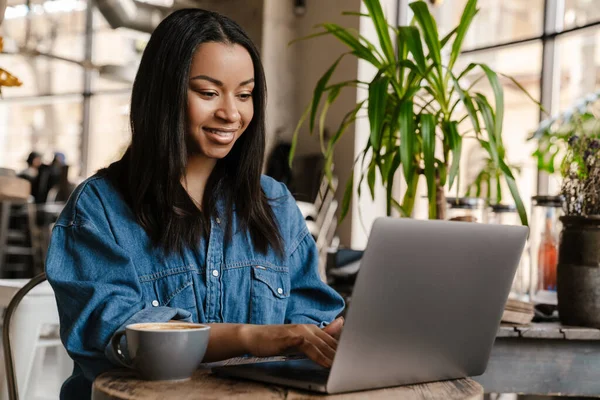 坐在咖啡桌旁 边喝咖啡边工作边学习 一边用手提电脑微笑着元朗的非洲女人 — 图库照片