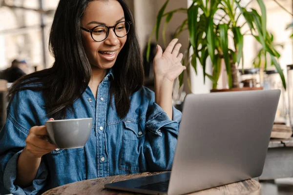 微笑着年轻的非洲女人 一边用手提电脑 一边坐在茶几边喝咖啡 一边工作 学习或打电话 一边挥手 — 图库照片