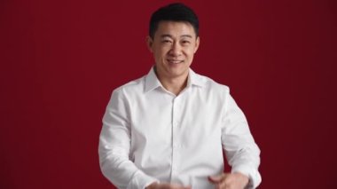 Beyaz tişörtlü gülümseyen Asyalı adam kırmızı stüdyodaki kameraya kalp şeklini gösteriyor.