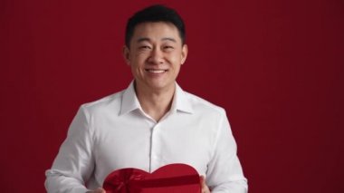 Beyaz gömlekli mutlu Asyalı adam kırmızı stüdyodaki kameraya hediye kutusu veriyor.