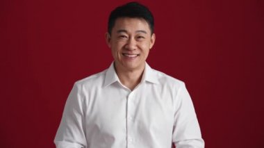 Beyaz gömlekli pozitif Asyalı adam kırmızı stüdyodaki kameraya hediye kutusu veriyor.