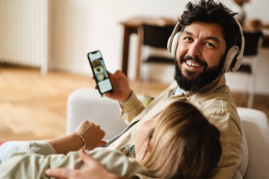 Beyaz çift evdeki koltukta otururken cep telefonu ve kulaklık kullanıyor.