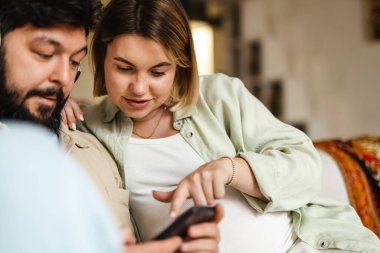 Beyaz çift evdeki koltukta otururken cep telefonu ve kulaklık kullanıyor.