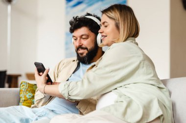 Beyaz çift evdeki koltukta otururken cep telefonu ve kulaklık kullanıyor.