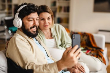Beyaz çift evdeki koltukta otururken cep telefonu ve kulaklık kullanıyor.
