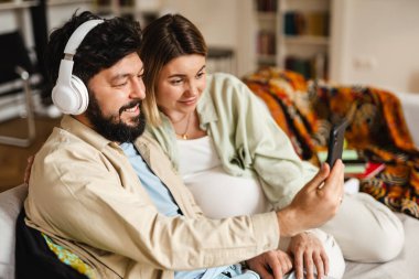 Beyaz çift evdeki koltukta otururken cep telefonu ve kulaklık kullanıyor.