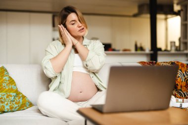 Genç hamile bir kadın evdeki kanepede otururken el kol hareketi yapıp dizüstü bilgisayar kullanıyor.